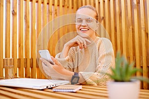 Image of attractive young adult woman with bun hairstyle wearing beige sweater sitting at table with closed laptop and holding