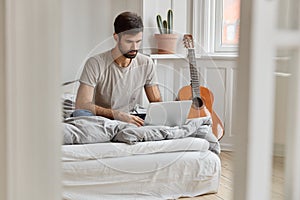 Image of attractive young male sits in comfortable bed, synchronizes multimedia files on laptop computer, works