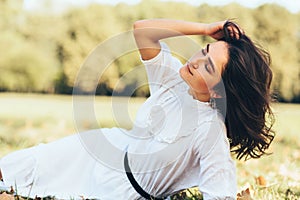 Image of attractive young woman with windy hair lying on the grass, enjoying the warm weather, wearing white dress on nature