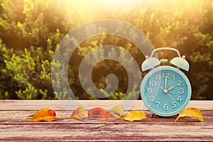 Image of autumn Time Change. Fall back concept. Dry leaves and vintage alarm Clock on wooden table outdoors at afternoon