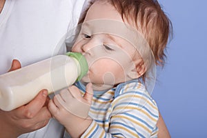 Image of baby drinking milk and looking at his bottle, isolated over blue background, wearing striped shirt, feels happyness in