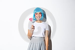 Image of beautiful asian girl in blue short wig celebrating halloween, holding candy and smiling at camera, standing