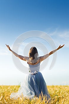 Image of beautiful blond young woman wearing long blue ball dress with arms wide expand looking up on wheat field and blue sky photo