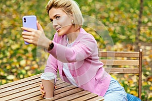 Image of beautiful blonde young woman wearing blue jeans, white t-shirt and pink jacket, smiling and taking selfie on smartphone.