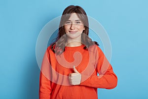 Image of beautiful brunette woman wearing casual clothes showing thumbs up and looking directly at camera at camera with cute