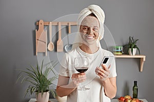 Image of beautiful Caucasian woman with cosmetic mask in white towel wrapped around head drinking wine in kitchen, looking at