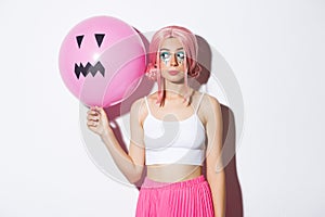 Image of beautiful glamour girl in pink wig, bright makeup, holding balloon with scary halloween face, standing over
