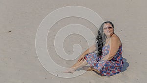 Image of a beautiful happy and smiling woman sitting on the sand in a blue dress with red and white flowers
