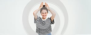 Image of beautiful, happy young woman showing bunny ears on top of her head, looking excited, posing over white