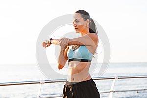 Image of beautiful healthy woman 20s in sports bra looking forward, and stretching arms near seaside in sunny morning