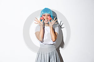 Image of beautiful smiling asian girl looking silly, showing macaroons, wearing blue anime wig, standing over white