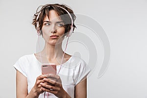 Image of beautiful woman in basic t-shirt holding smartphone while listening to music with headphones