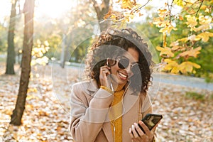 Beautiful young african happy woman walking outdoors in a spring park using mobile phone listening music with earphones