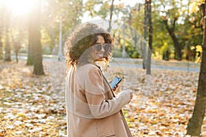 Beautiful young african happy woman walking outdoors in a spring park using mobile phone listening music with earphones