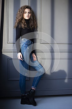Image of a beautiful young woman with curly hair, in black suitcase and jeans, posing near grey wall.