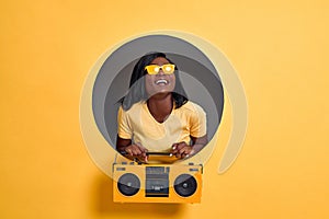 Image of beautiful young woman in yellow sunglasses laughing and holding vintage boombox isolated over circle hole in yellow