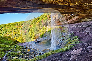 Behind huge waterfall tucked into cliffs with open view of fall forest mountains and small tourists below