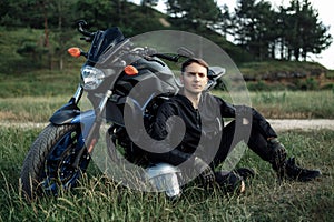 Image of biker sitting on the grass in front of motorcycle in sunset on the country road.