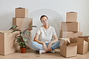 Image of bored tired woman wearing white t shirt sitting on floor surrounded with cardboard boxes with belongings, being exhausted