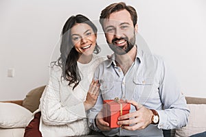 Image of brunette couple holding birthday present box while sitting on sofa in bright room