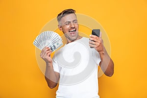 Image of brunette guy 30s in white t-shirt holding cell phone and bunch of money