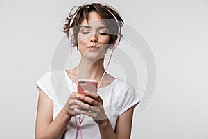 Image of brunette woman in basic t-shirt holding smartphone while listening to music with headphones