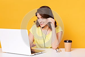 Image of brunette woman in yellow casual t shirt sits on chair at white deskand working on laptop over studio background, has