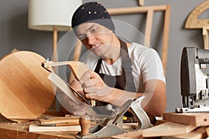 Image of calm attractive young adult man carpenter wearing white t-shirt, black cap and brown apron working in joinery, making