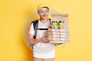 Image of calm smiling deliveryman with thermo backpack in white T-shirt and cap standing against yellow wall, holding and looking
