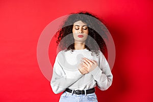 Image of calm young woman with curly hairsty, close eyes and holding hands on heart, keeping warm memories, feeling