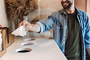 Image of caucasian cheerful man throwing paper garbage to separate collection bins for recycling in cafe indoors