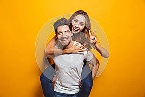 Image of caucasian couple having fun while man piggybacking joyful woman, isolated over yellow background