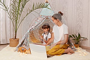 Image of Caucasian dark haired woman sitting on floor near children`s teepee with her daughter, little girl using mobile phone,