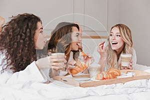 Image of caucasian smiling women 20s wearing white housecoat having breakfast in bed in luxuty apartment or hotel room, during ba photo