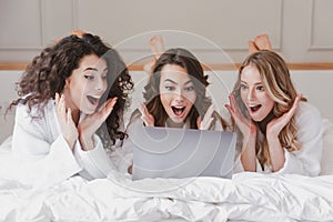 Image of caucasian smiling women 20s wearing white housecoat lying in luxury bedroom at home, and using silver laptop during bach photo
