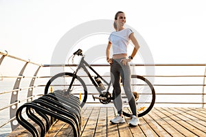 Image of caucasian woman standing with bicycle on boardwalk, during sunrise over sea