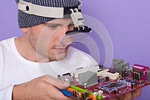 Image of Caucasian young technician using magnifying glass for repairing personal computer`s motherboard in his lab. Concept of