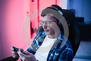 Image of caucasian youngman in headphones using cellphone playing video game in a gaming chair at home