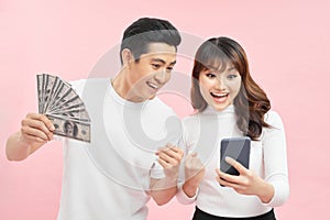 Image of cheerful young asian loving couple standing isolated over pink wall background holding money and mobile phone