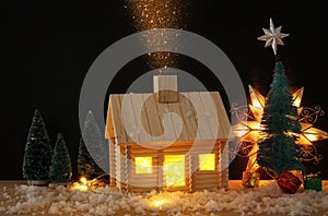 Image of christmas tree and wooden house with light through the window, over snowy table.