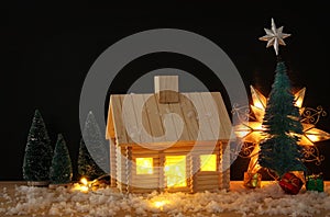 Image of christmas tree and wooden house with light through the window, over snowy table.