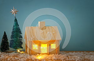 Image of christmas tree and wooden house with light through the window, over snowy table.