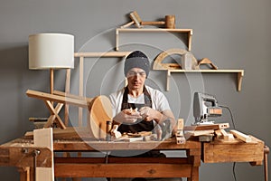 Image of concentrated man carpenter wearing brown apron and black cap making a handmade wooden toy in a home workshop. enjoying