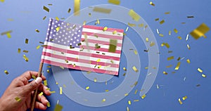 Image of confetti falling over hand holding flag of united states of america on blue background