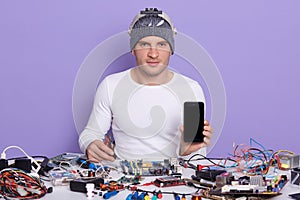 Image of confident professional radiotrician sitting at his workplace, holding smartphone and side cutting pliers, wearing grey