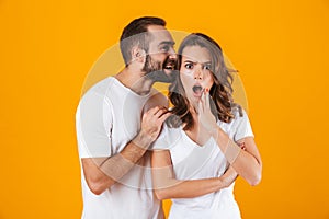 Image of content man whispering secret or interesting gossip to woman in her ear, isolated over yellow background