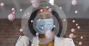 Image of covid 19 cells floating over portrait of biracial woman wearing face mask in office