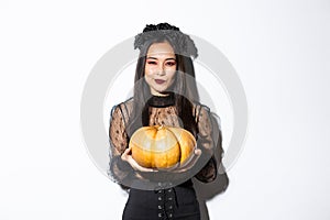 Image of cunning asian woman in black dress, impersonating evil witch on halloween, holding big pumpkin, standing over