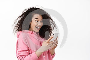 Image of curly girl look surprised at mobile phone screen, receive exciting news message on smartphone, chatting in app