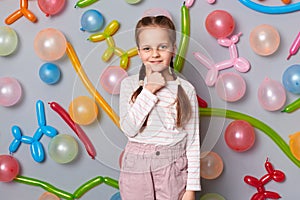 Image of cute charming smiling little girl with braids wearing casual clothing posing isolated over gray background with balloons